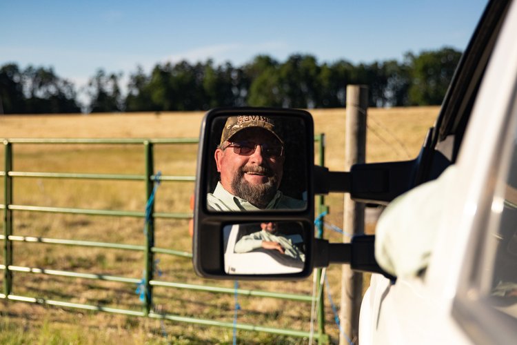 A smiling man wearing sunglasses and a camouflage hat is reflected in the side mirror of a vehicle, with a rural landscape and a green fence in the background.
