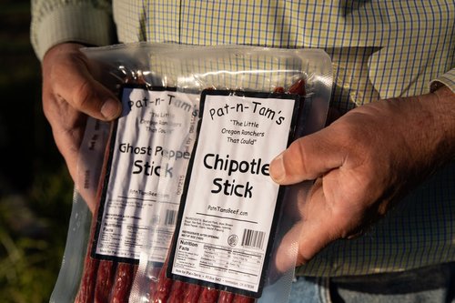 Person holding two packages of Pat-n-Tam's meat sticks, featuring Ghost Pepper Stick and Chipotle Stick flavors, outdoors with a blurred background.