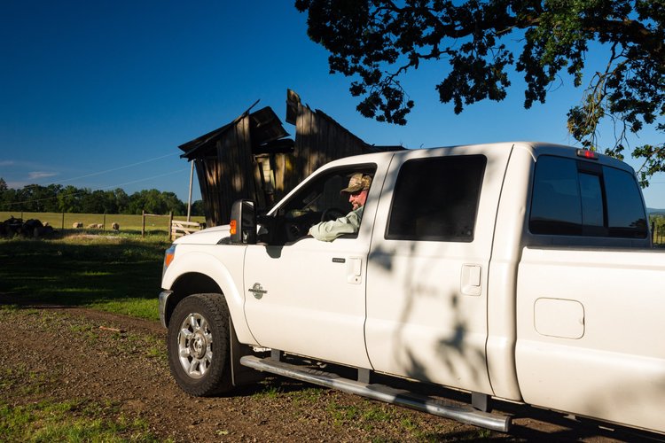 A man wearing a camouflage hat sits in the driver's seat of a white pickup truck, parked near an old barn in a rural setting with green fields and a clear blue sky in the background.