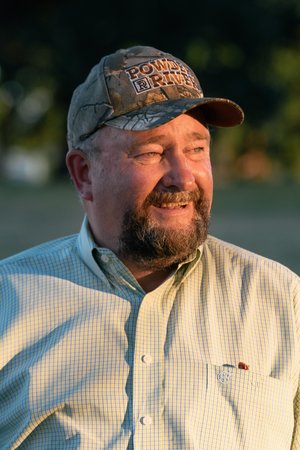 Man wearing a camo hat and light green checkered shirt, smiling outdoors in a natural setting during sunset.