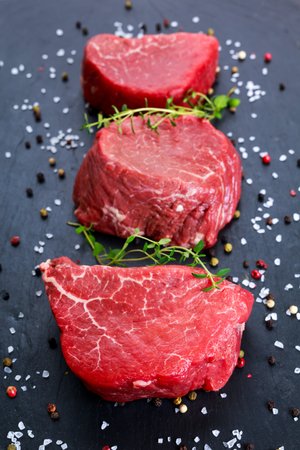 Three cuts of fresh red meat arranged on a dark slate surface, garnished with fresh thyme and surrounded by colorful peppercorns and sea salt.