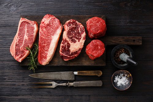 Raw cuts of premium beef including ribeye, filet mignon, and sirloin displayed on a wooden cutting board, accompanied by kitchen tools, a mortar and pestle with spices, and coarse salt.