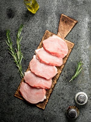 Raw pork chops arranged on a wooden cutting board, garnished with fresh rosemary sprigs and surrounded by olive oil and salt and pepper shakers, set against a textured gray background.