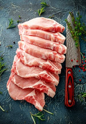 Freshly cut pork chops arranged neatly on a textured surface, accompanied by a sharp chef's knife and garnished with herbs. Ideal for cooking and grilling recipes.