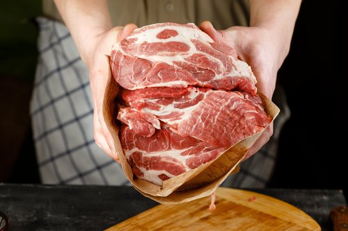 Fresh cuts of marbled pork shoulder being held in brown butcher paper, ready for cooking or preparation in a kitchen setting.